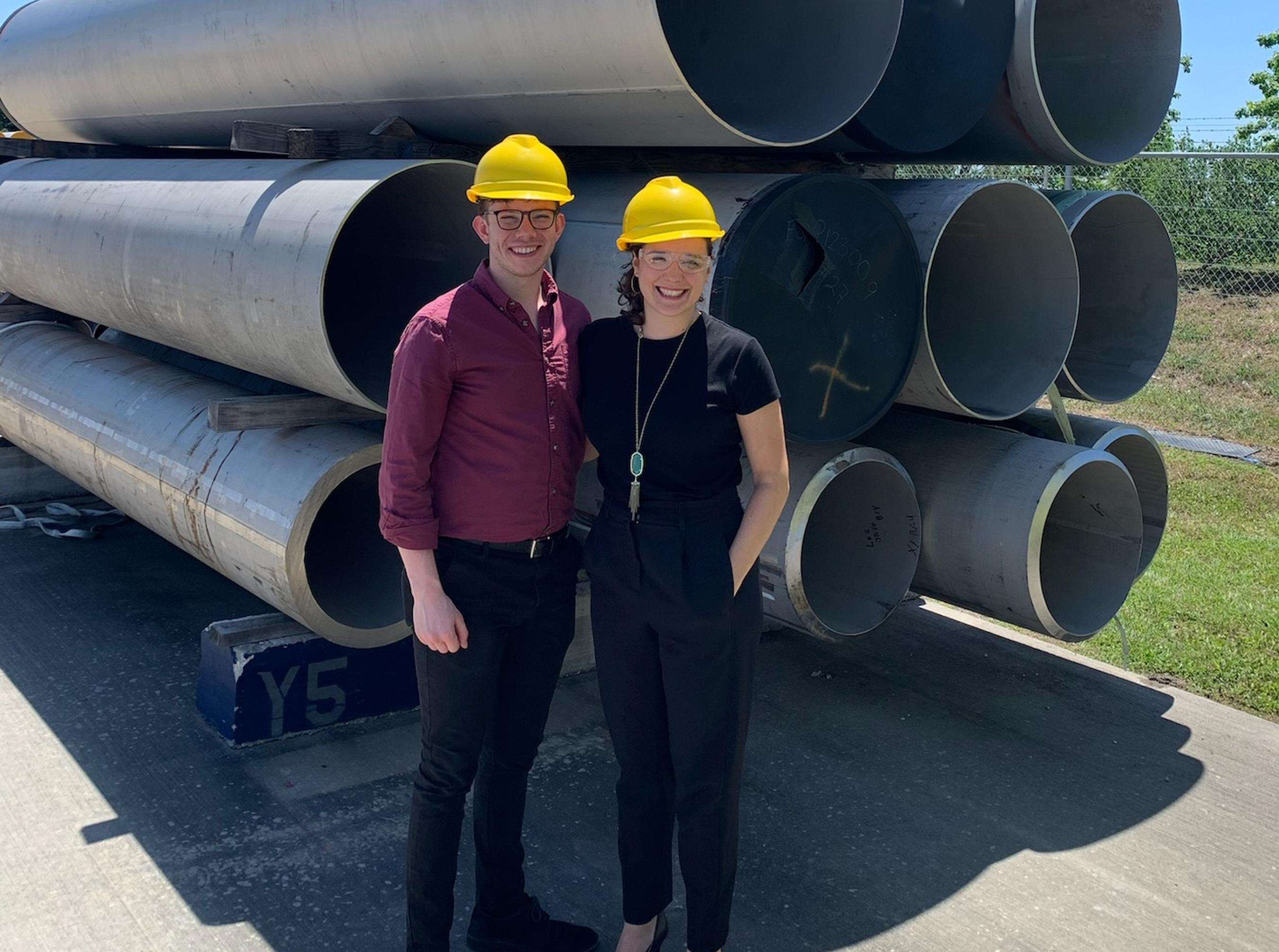 Two workers wearing yellow hardhats standing in front of steel pipes and smiling.